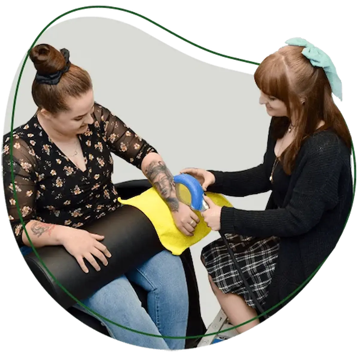 Two women, one receiving a treatment on her tattooed arm from the other using a blue and silver handheld device.