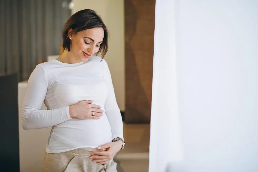 A smiling pregnant woman in a white top gently holds her belly with both hands, looking down.