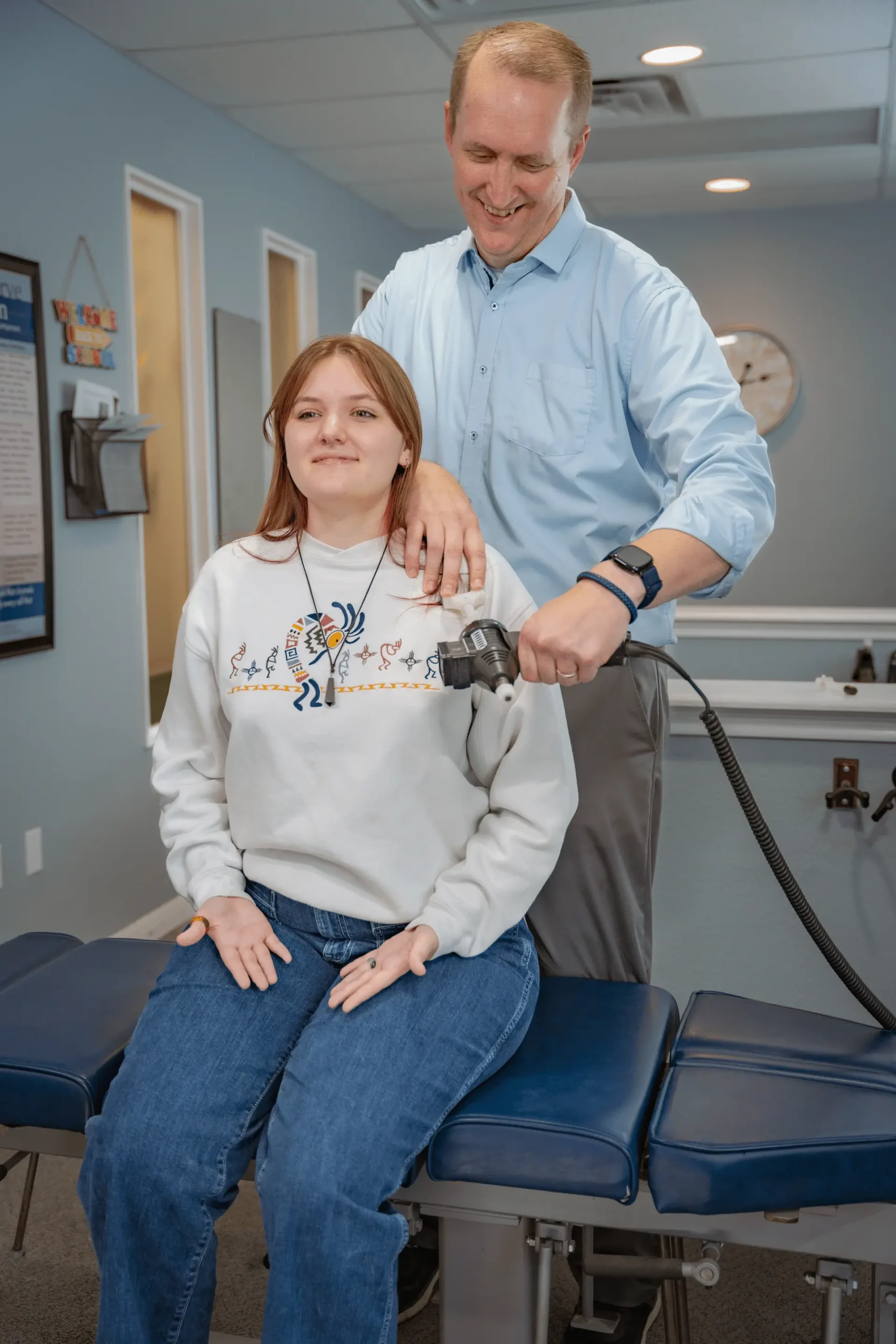 A male chiropractor uses a handheld adjustment tool on a female patient's upper back as she sits on a blue examination table.