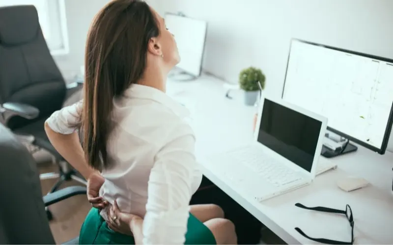 A woman in a white shirt and green skirt sits at a desk, holding her lower back and tilting her head back in discomfort.