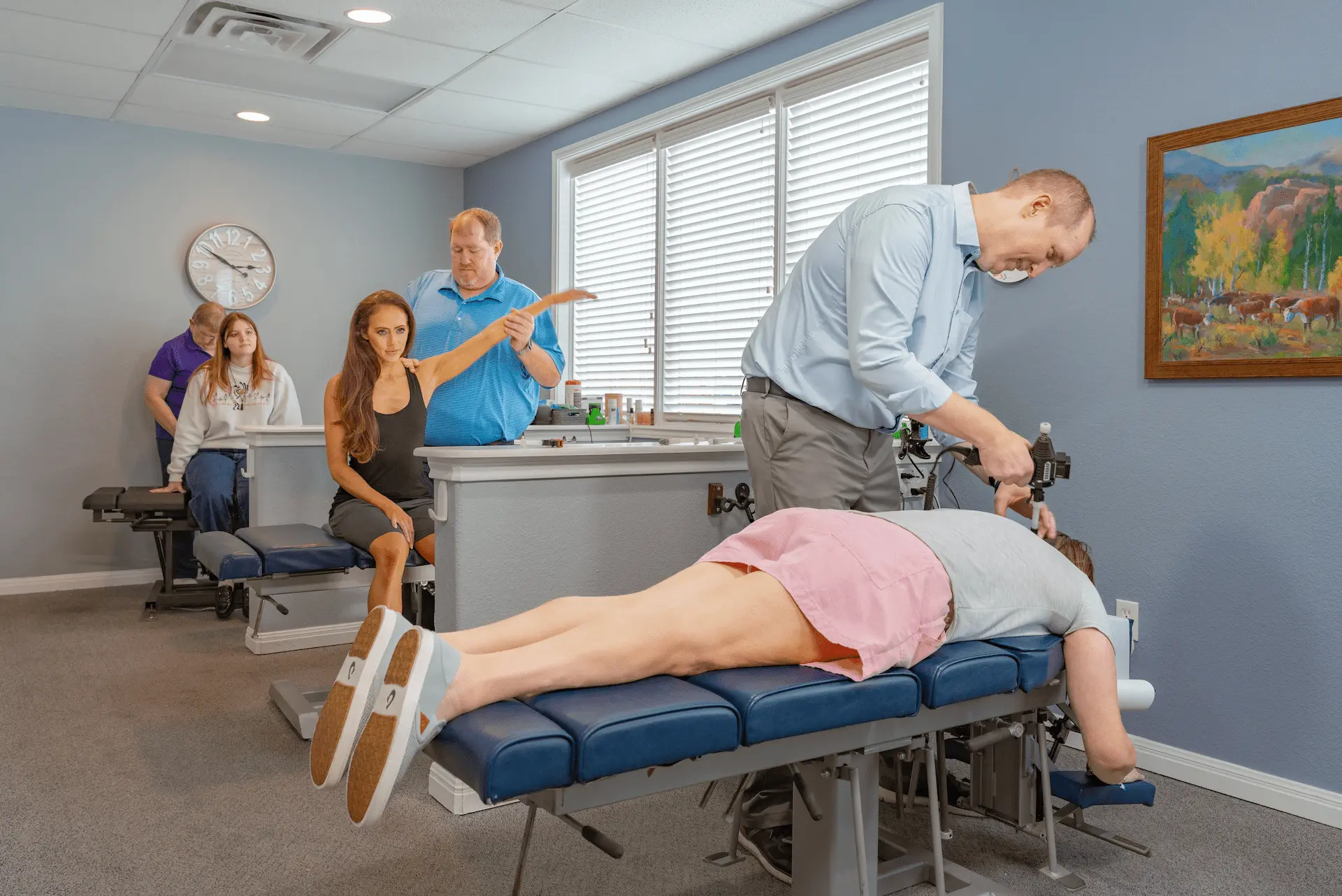 A chiropractic clinic with several patients and practitioners. In the foreground, a woman lies face down on a blue table while a male chiropractor uses an adjustment tool on her back. Other patients are seated, receiving assessments or adjustments from other practitioners.