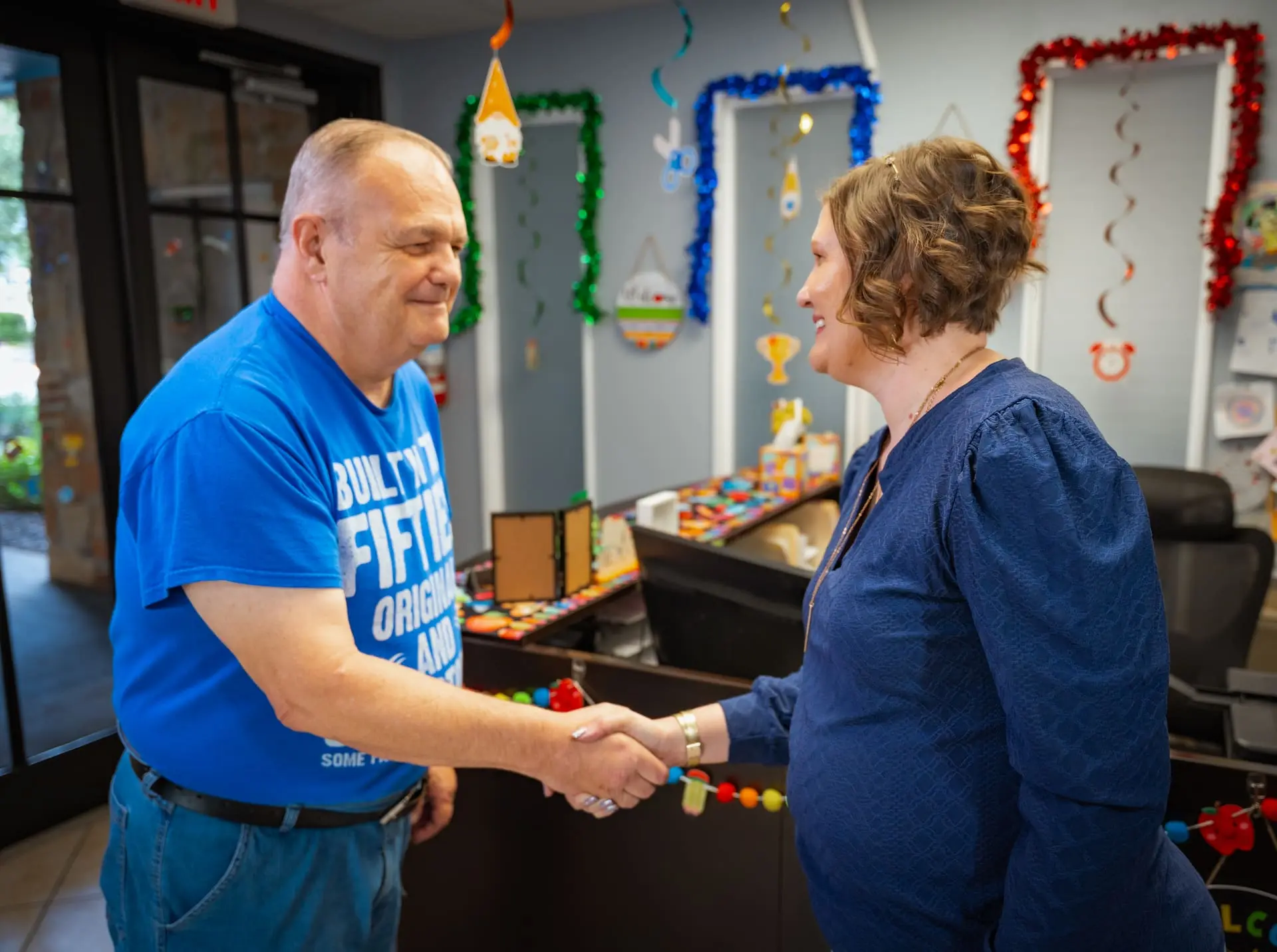 A man and a woman smiling and shaking hands in a colorful, decorated room.