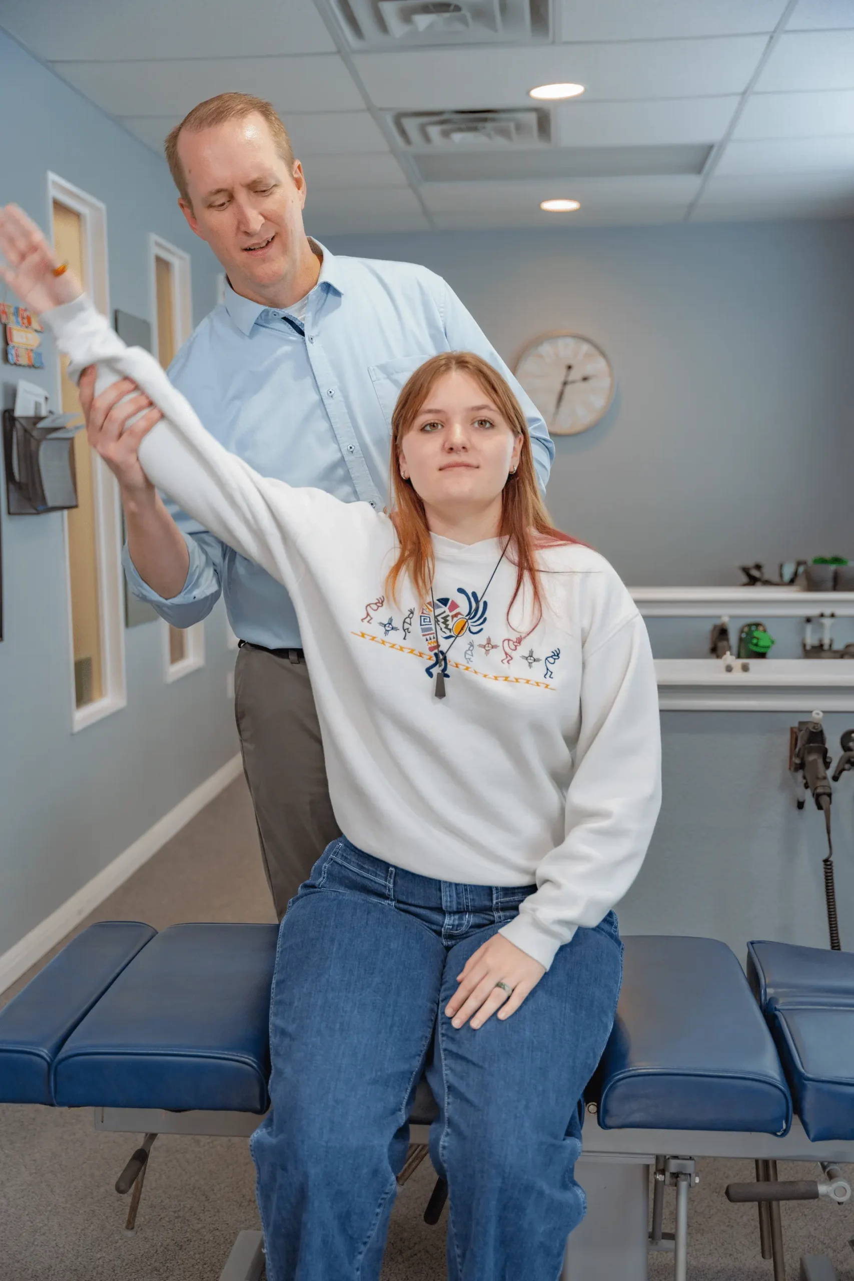A man in a light blue shirt helps a young woman in a white patterned sweatshirt raise her arm while she sits on a blue examination table in a medical office.