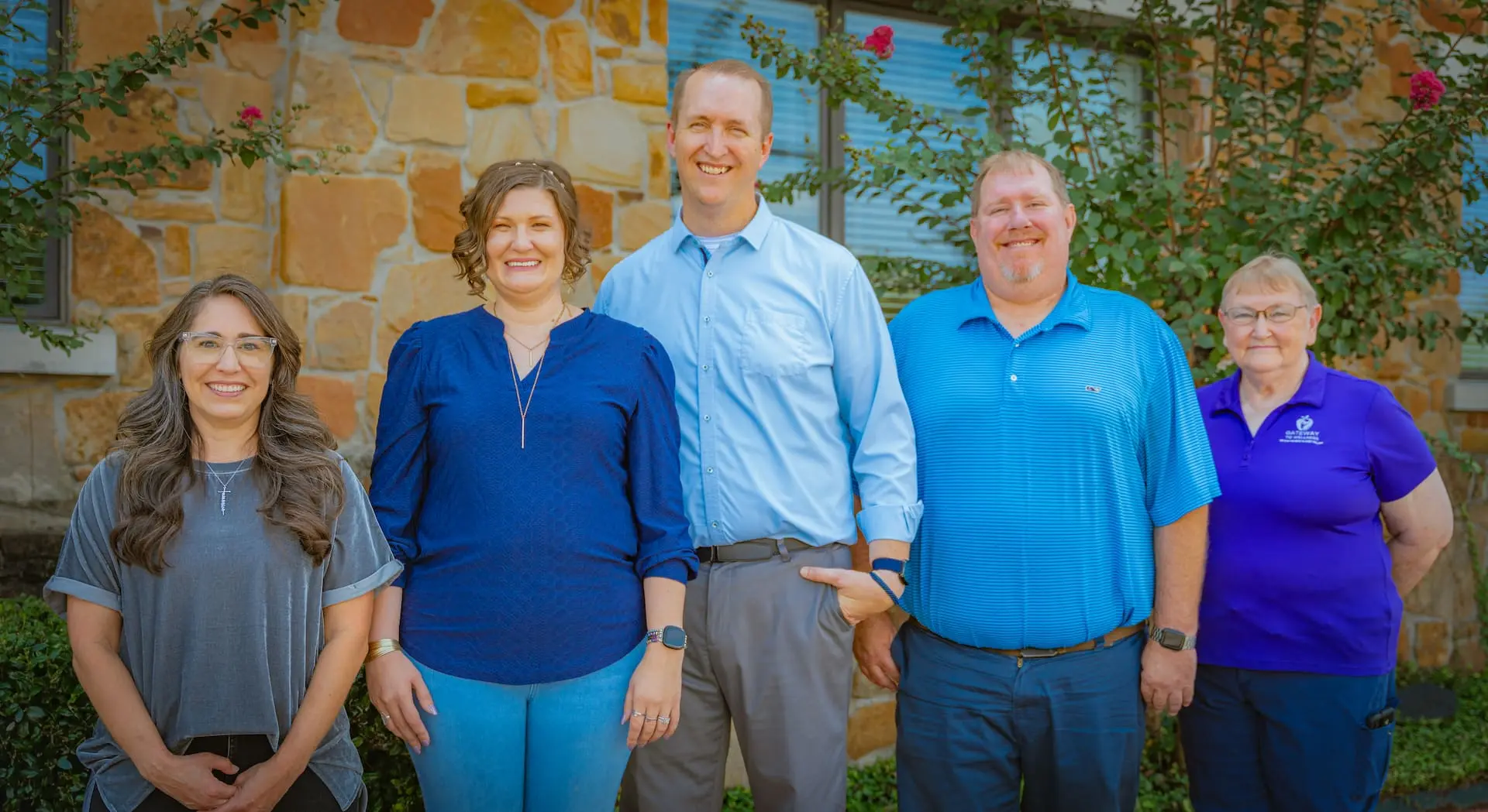 Five people, three women and two men, smiling and standing in a line outdoors in front of a stone building with green plants.
