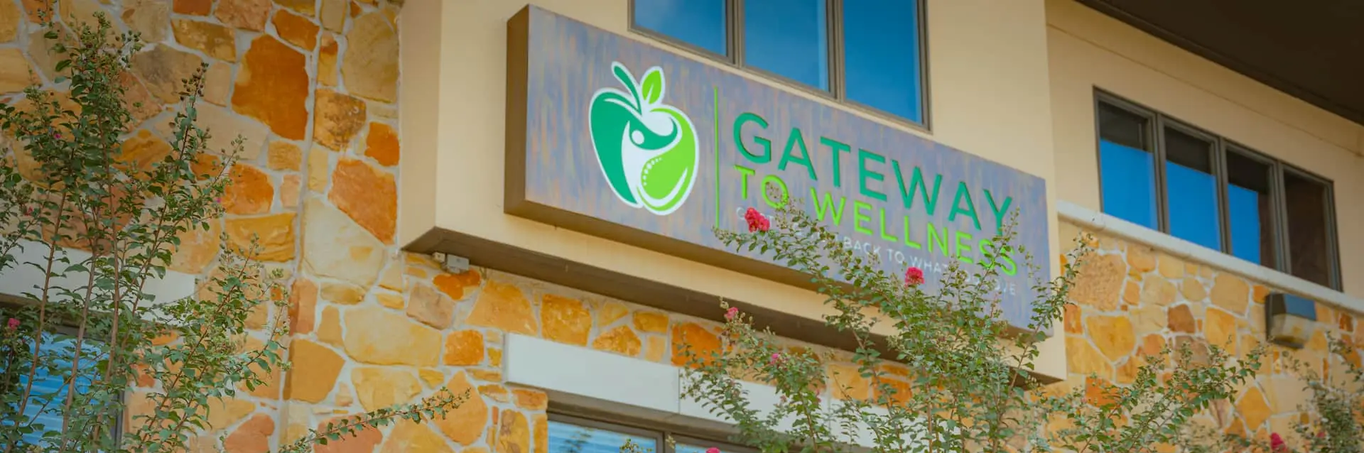 Exterior of a building with a stone facade and a sign that reads 'Gateway to Wellness' with a green apple logo. Green bushes with pink flowers are in the foreground.