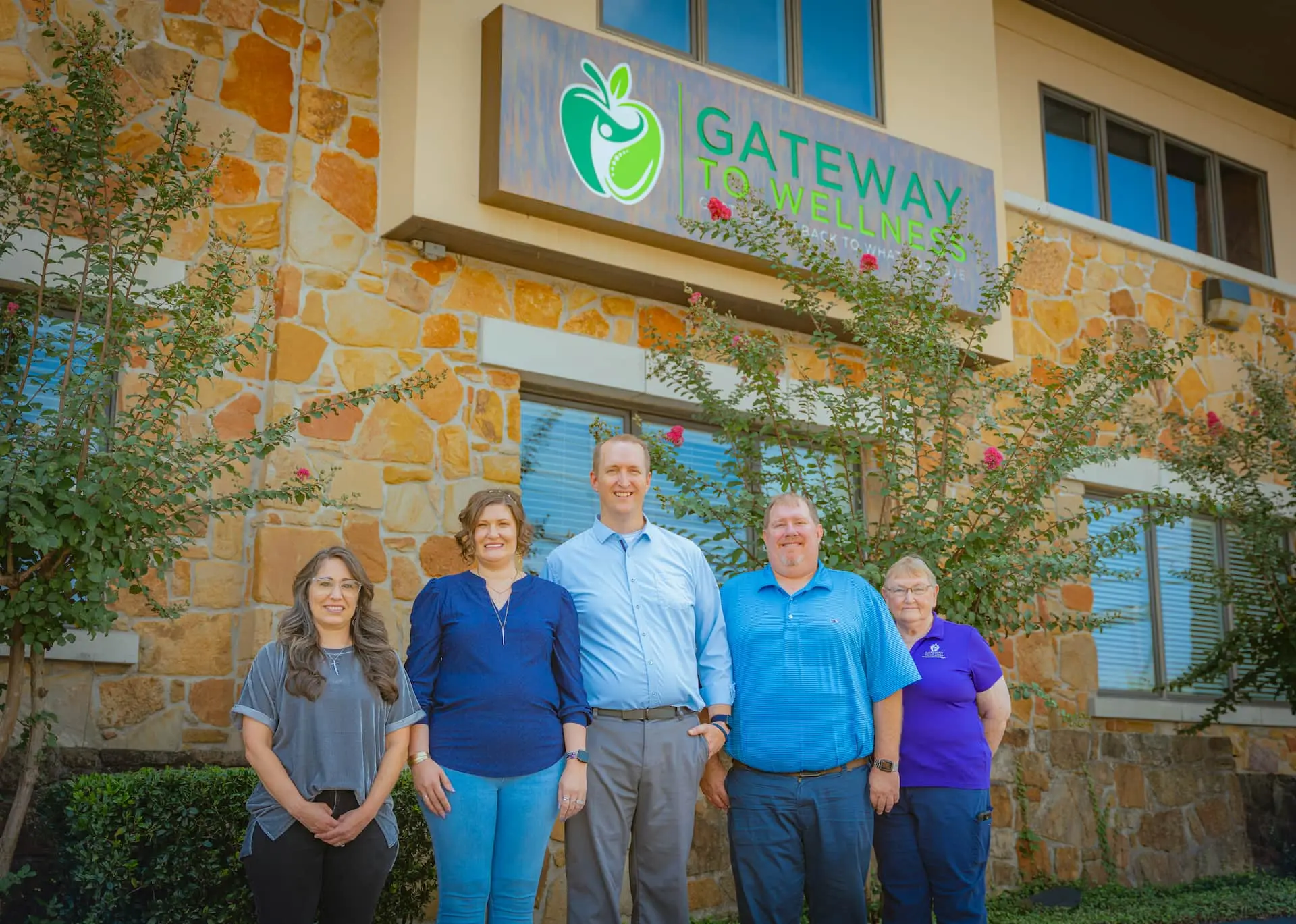 Five team members, three women and two men, smiling in front of the Gateway to Wellness building with its green apple logo.