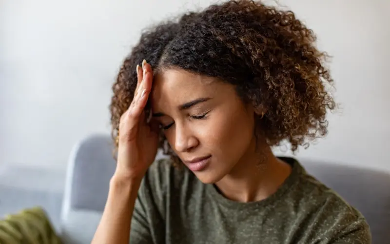 A young woman with curly hair holds her hand to her forehead, eyes closed, looking distressed.