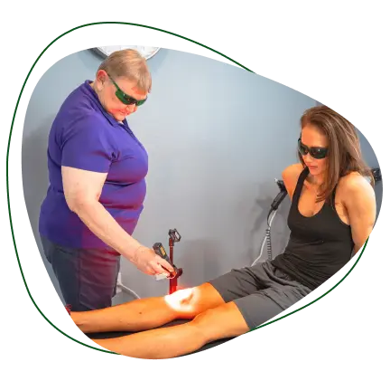 A woman in a purple shirt and green glasses administers red light therapy to another woman's knee, who is seated and wearing dark glasses.