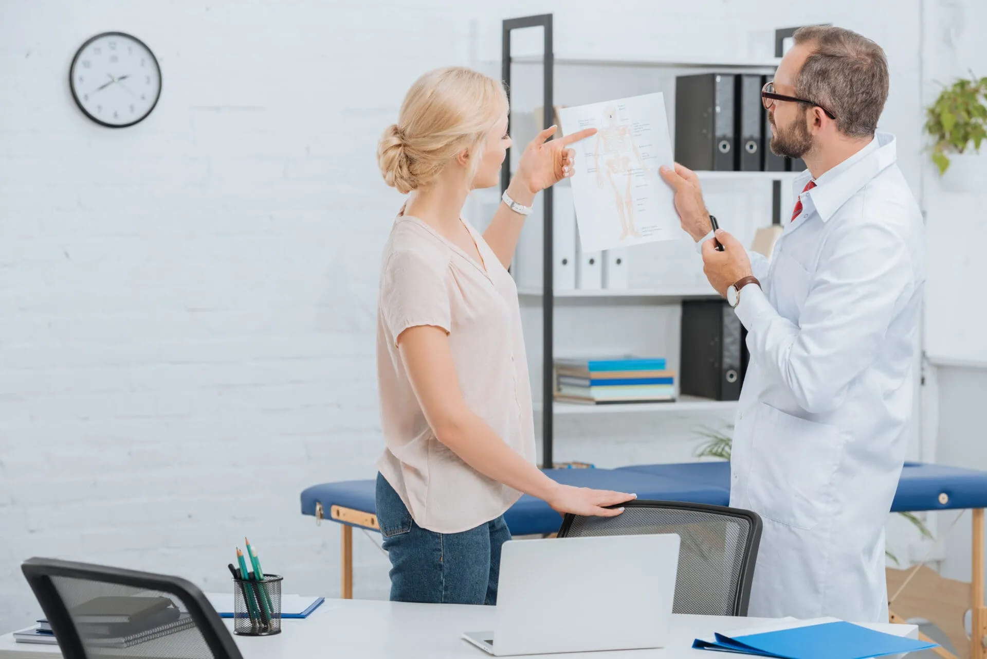 A doctor and a patient standing, discussing an anatomical chart in a medical office.