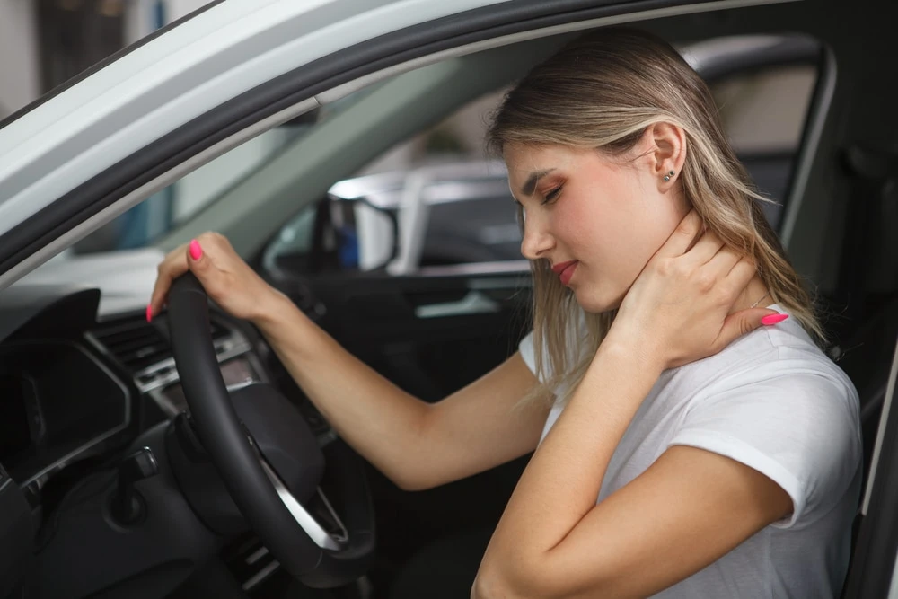 A woman in a white t-shirt sits in a car, holding the back of her neck with her right hand, appearing to be in pain.