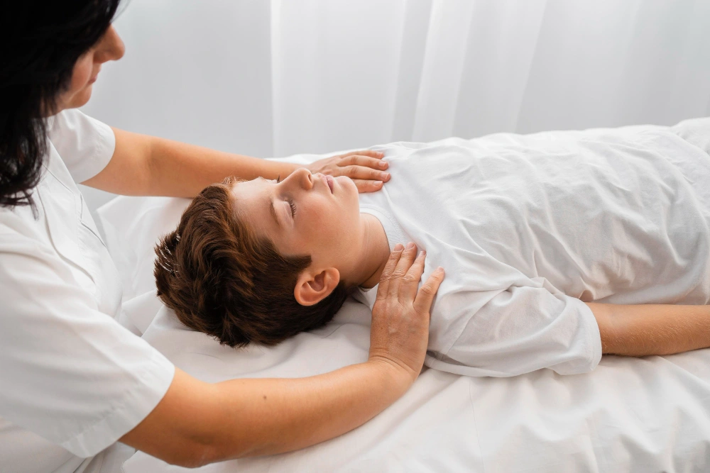 A woman in a white uniform gently places her hands on a child lying on their back with eyes closed, on a white bed.