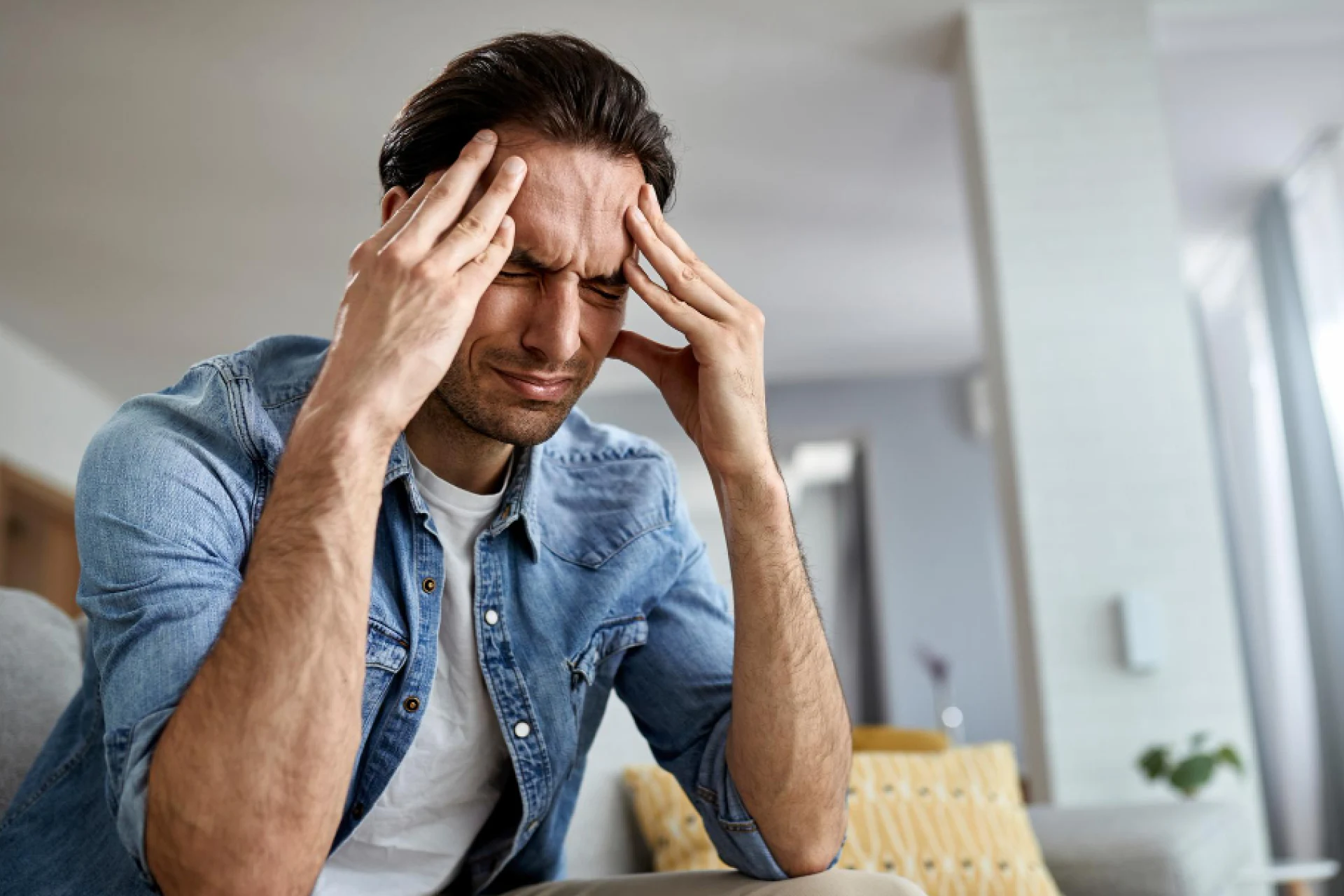 A man with dark hair, wearing a denim shirt, sits with his eyes closed and hands pressed to his temples, indicating a headache or stress.