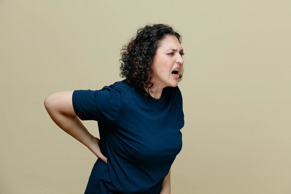 A woman in a blue t-shirt holds her lower back and screams, indicating pain.