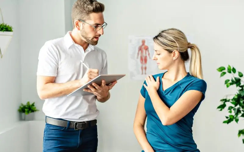 A male physiotherapist in glasses takes notes on a tablet while a female patient in a teal shirt holds her shoulder, indicating pain.