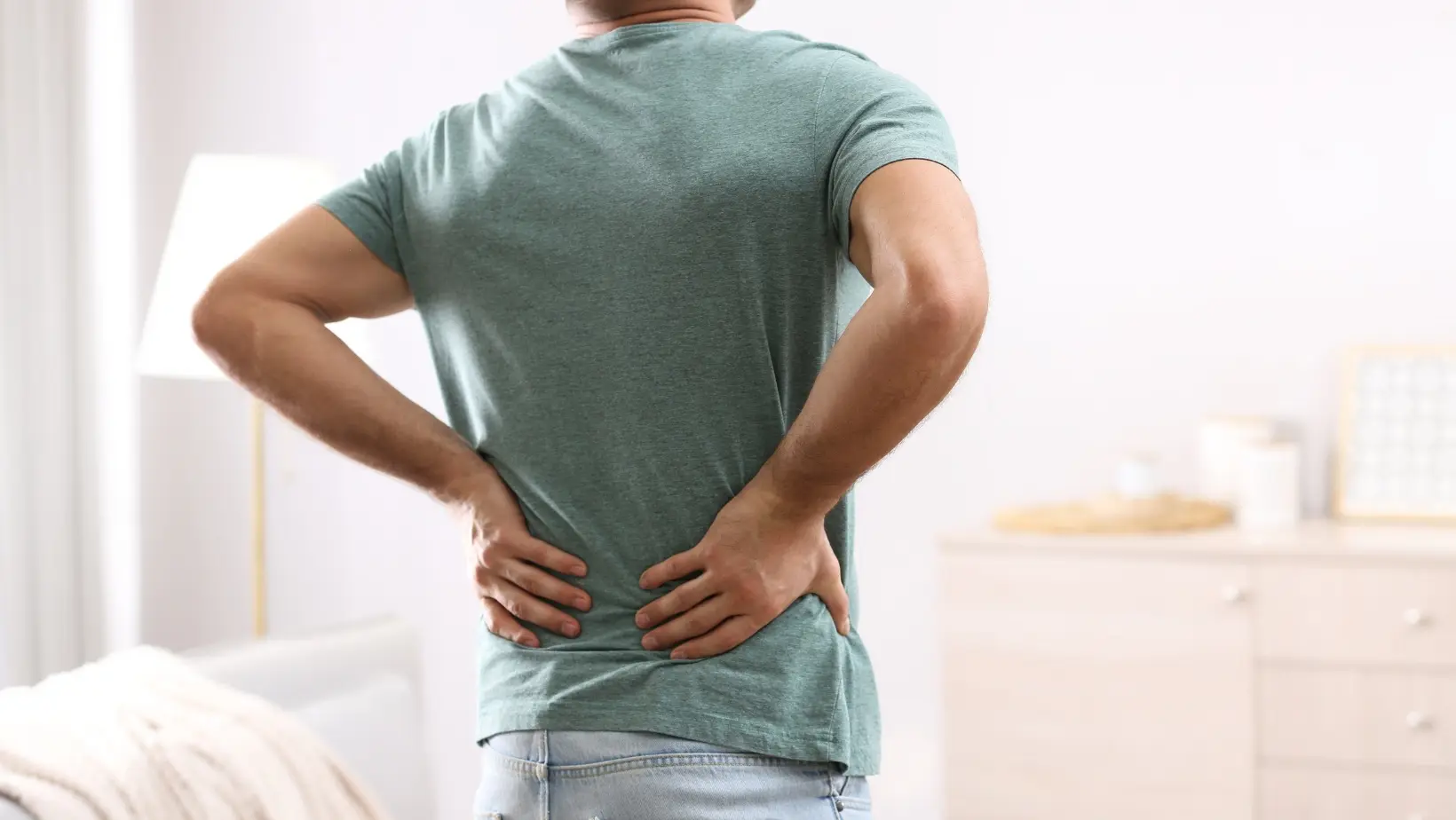Man with a blue t-shirt standing with his hands on his lower back, gesturing in pain. He is facing away from the camera.