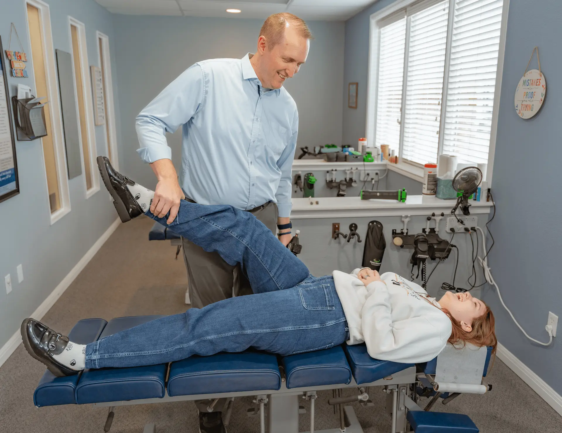 A male chiropractor adjusts a female patient's leg as she lies on a blue examination table.
