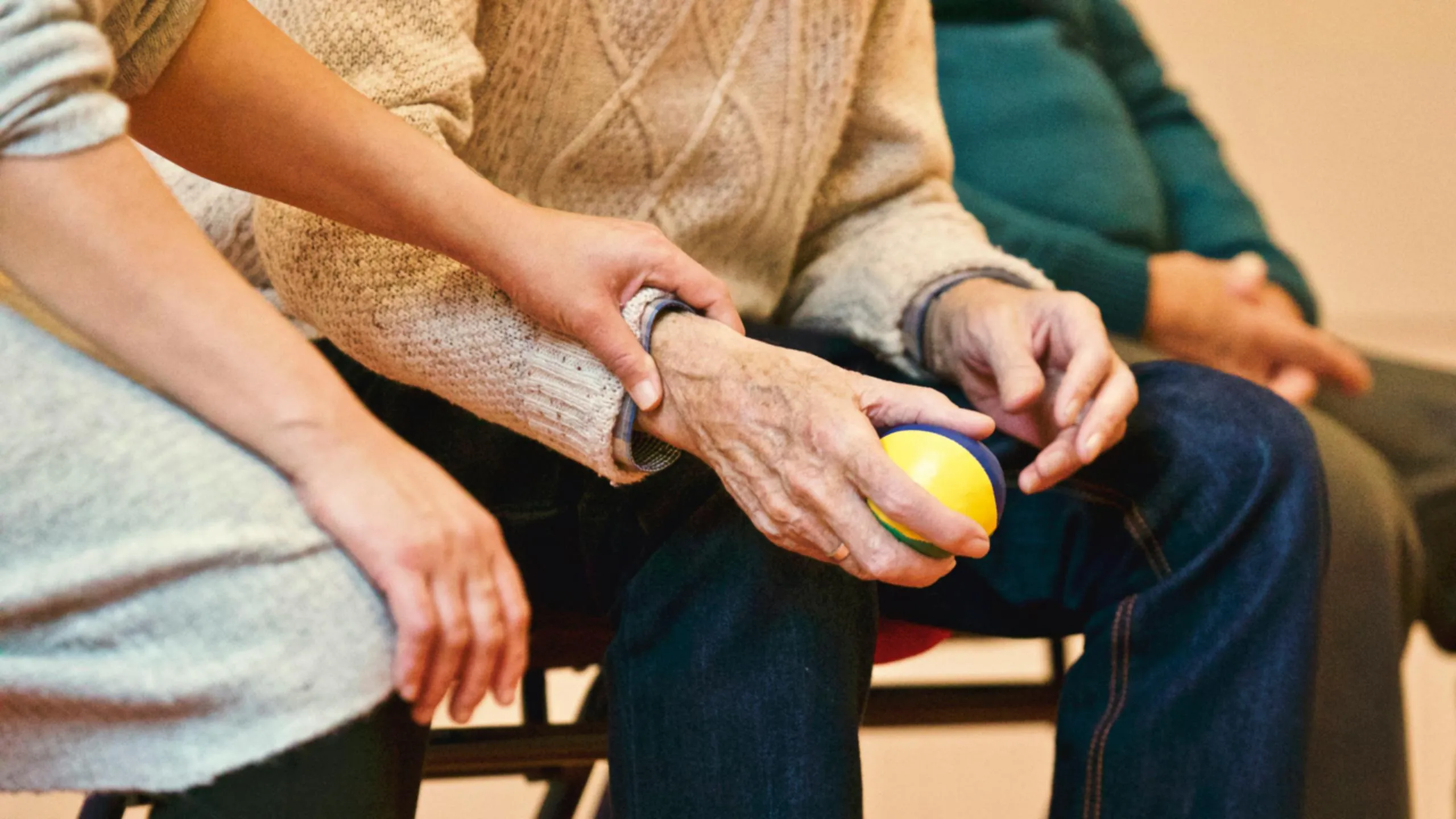 A younger person's hand gently holds the wrist of an older person, who is holding a yellow, blue, and green ball.