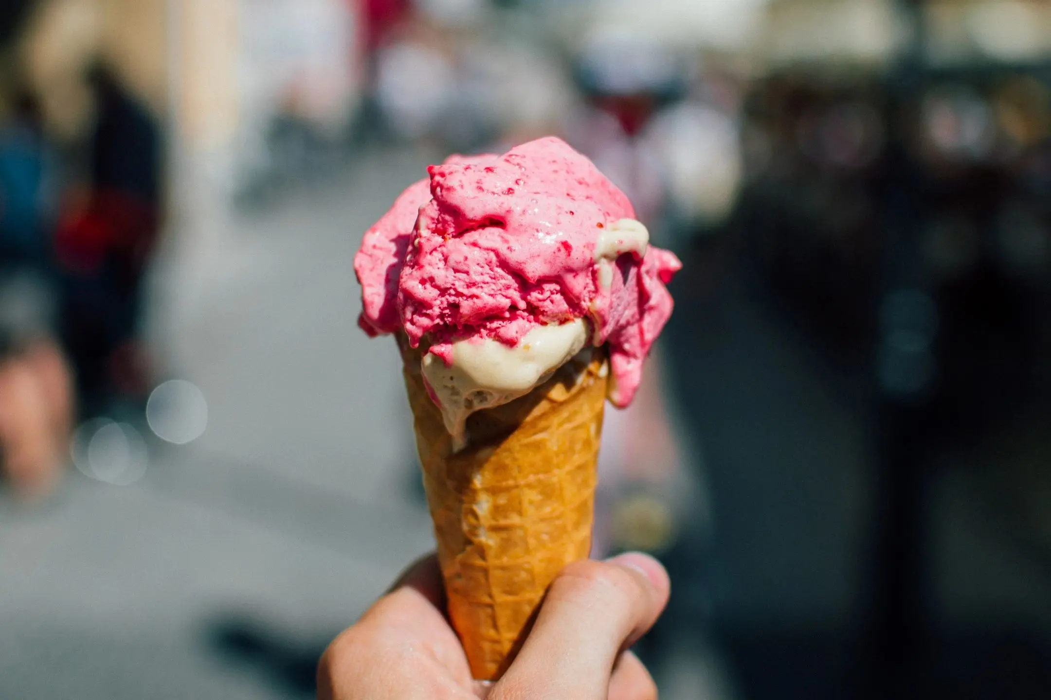 A hand holds a melting pink and beige ice cream cone on a sunny day.