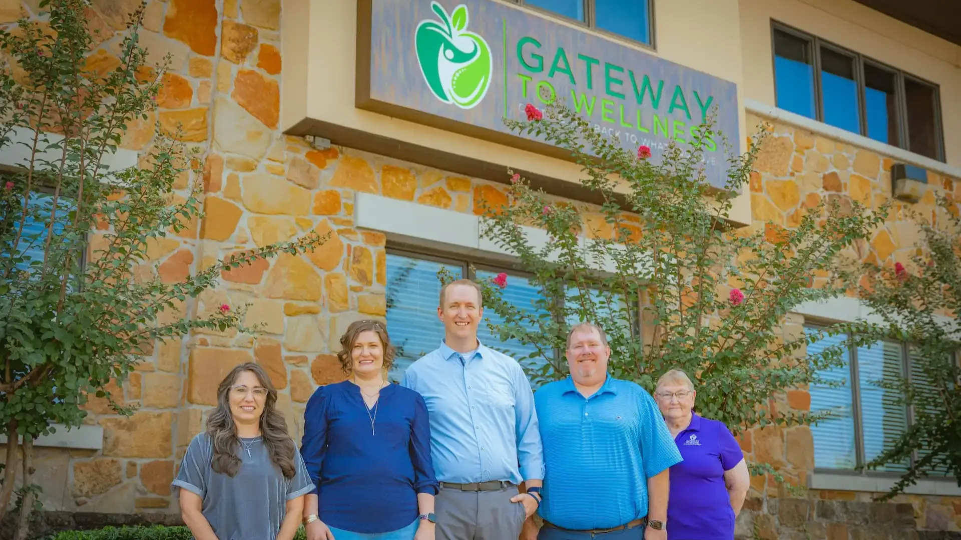 Five smiling people, three women and two men, stand in front of a building with a 'Gateway to Wellness' sign.