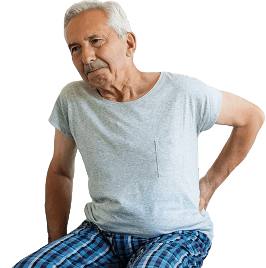 Elderly person facing in the direction of the camera, leaning forward with their hand on their back. Their expression suggests they are experiencing lower back and/or hip pain.