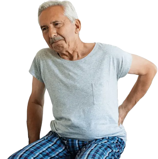 Elderly person facing in the direction of the camera, leaning forward with their hand on their back. Their expression suggests they are experiencing lower back and/or hip pain.