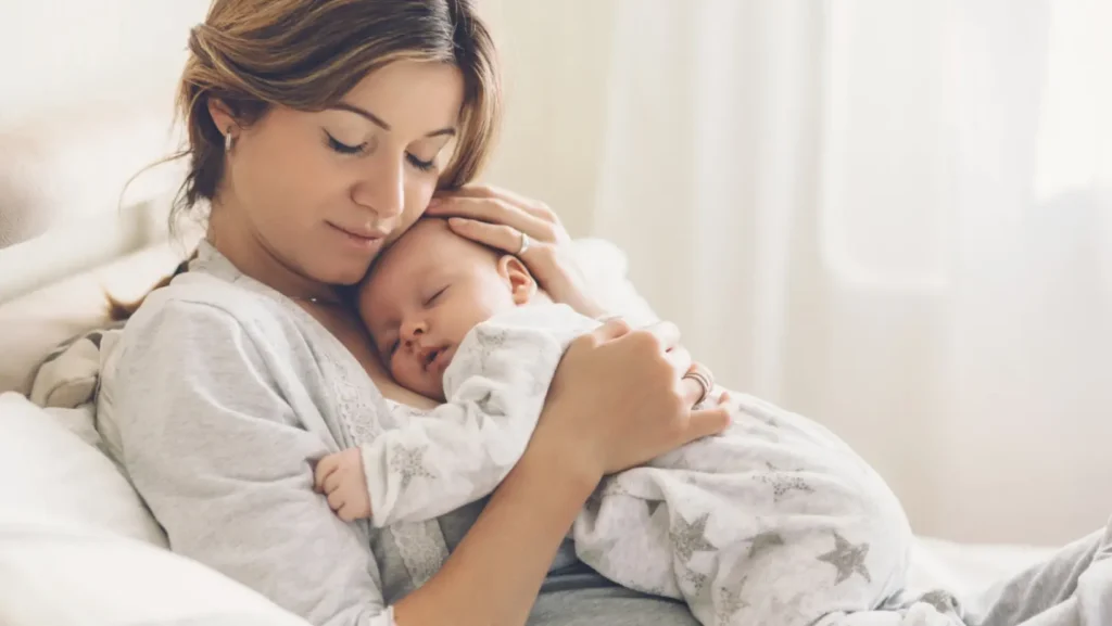 Featured Image for the Do I need postnatal care blog. Mother on couch with a sleeping baby held on her chest. The mother has her eyes closed, and is smiling with adoration.