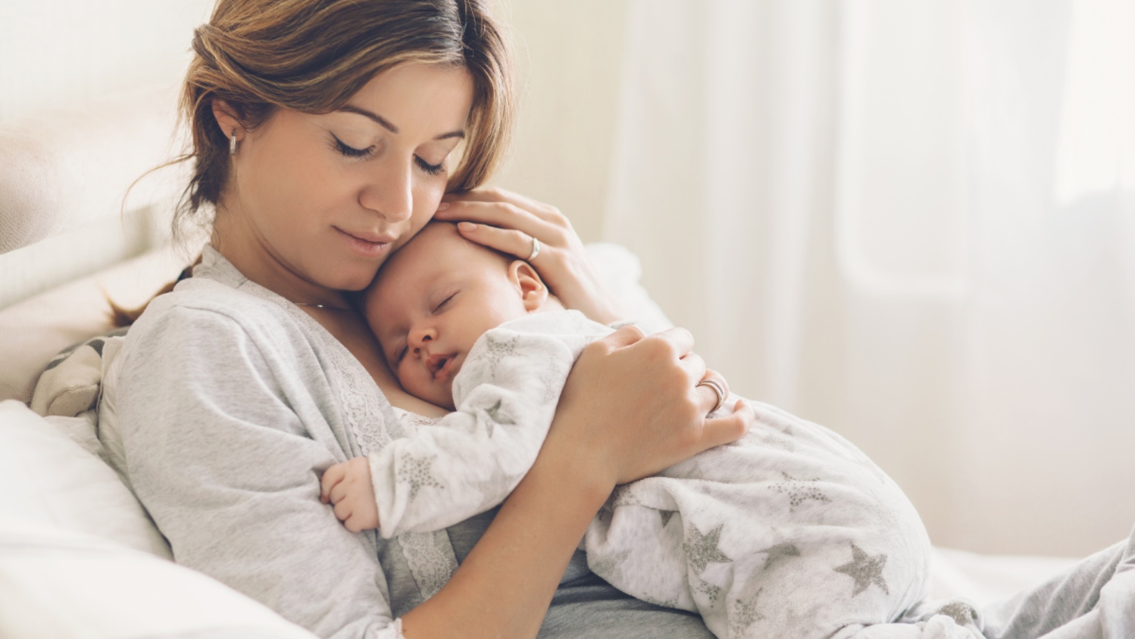 Featured Image for the Do I need postnatal care blog. Mother on couch with a sleeping baby held on her chest. The mother has her eyes closed, and is smiling with adoration.