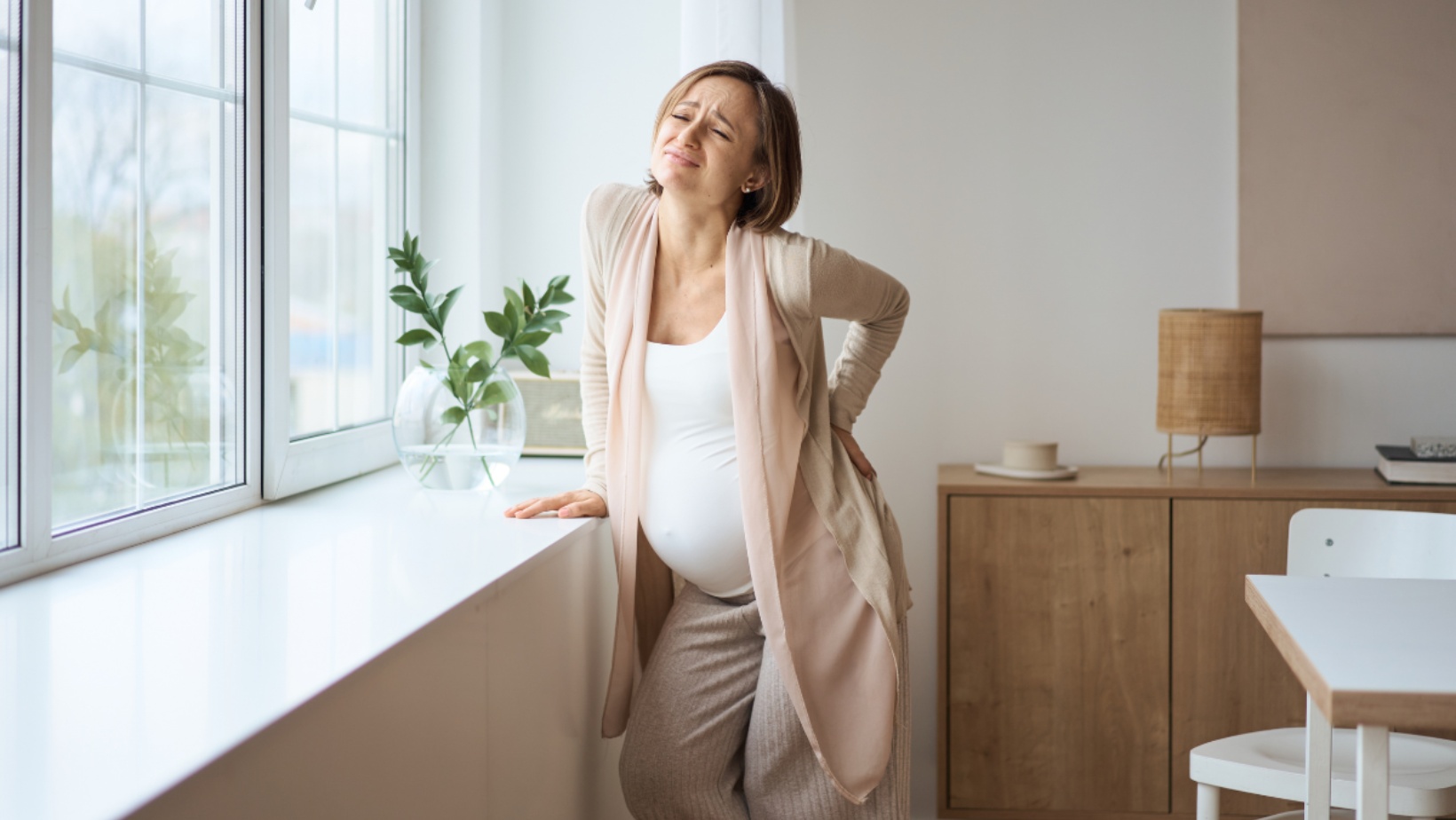 Pregnant person standing, leaning on a counter by a set of windows. They are expressing back pain, and their eyes are closed.
